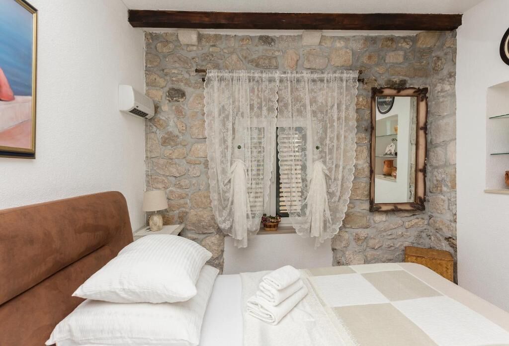 A bedroom with a stone wall, a bed with white linens, a brown headboard, a window with lace curtains, a mirror, and a wall-mounted air conditioner.