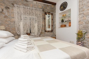 A room with a stone wall, a bed with a checkered bedspread, a stack of white towels, a mirror, and shelves with decorative items. - Apartman Listun (Makarska)