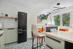 Cozinha moderna com geladeira preta, área de jantar com uma cadeira branca e vista para árvores através de grandes janelas.