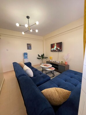 Ein Wohnzimmer mit einem blauen Sofa, einem kleinen runden Tisch, einem an der Wand montierten Fernseher und einer Deckenleuchte.