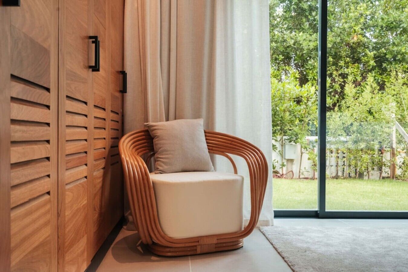 A wicker chair with a cushion in a room with a large window, wooden paneling, and a view of greenery outside.