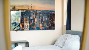 A hotel room with a bed, a nightstand, a painting of a cityscape, and a view of the city. - Peaceful 4-Bed Home in Aylesbury (Aylesbury)