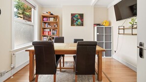 A dining area with a wooden table and four chairs, a bookshelf, a painting, a television, and a window with a view of trees. - Peaceful 4-Bed Home in Aylesbury (Aylesbury)