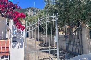 A gated entrance with a wrought iron gate, a white building with a balcony, and a red flowering plant. - Appartamento Tuttavista (Cala Gonone)