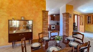 Un comedor con una mesa de madera dispuesta para cuatro personas, rodeada de sillas de madera. A la izquierda hay un aparador de vidrio y a la derecha, un pilar de ladrillo.
