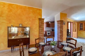 A dining area with a wooden table set for four, surrounded by wooden chairs. A glass display cabinet is to the left, and a brick pillar is to the right. - Appartamento Tuttavista (Cala Gonone)