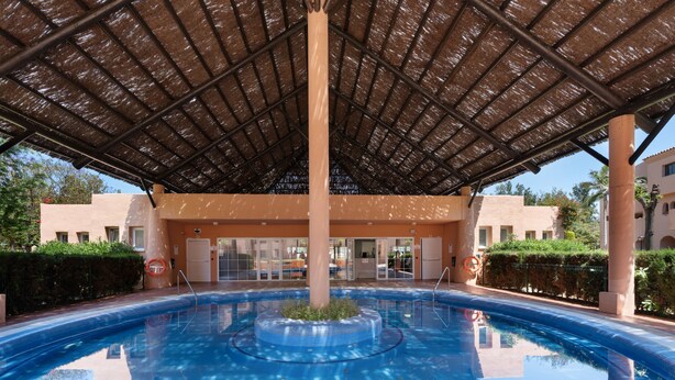 A swimming pool with a central island, a covered area, and a building with a beige facade in the background. - 'Inner Paradise' Apartment with Shared Pool, Wi-Fi and Air Conditioning (Atalaya Isdabe)