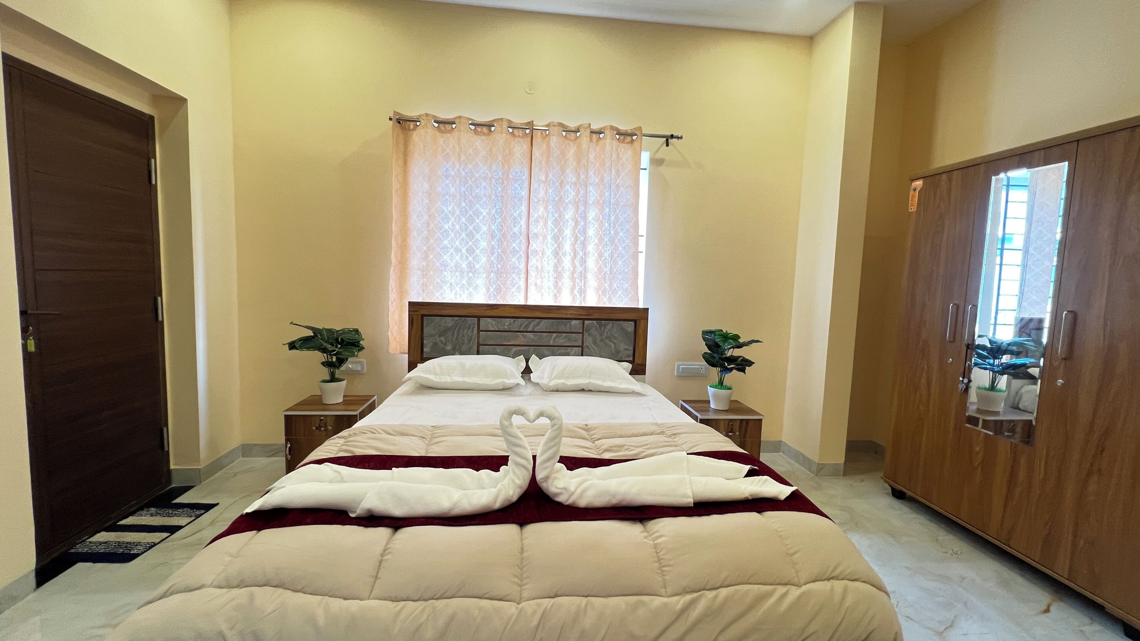A hotel room with a bed, two bedside tables with plants, a wooden wardrobe, and a window with curtains.