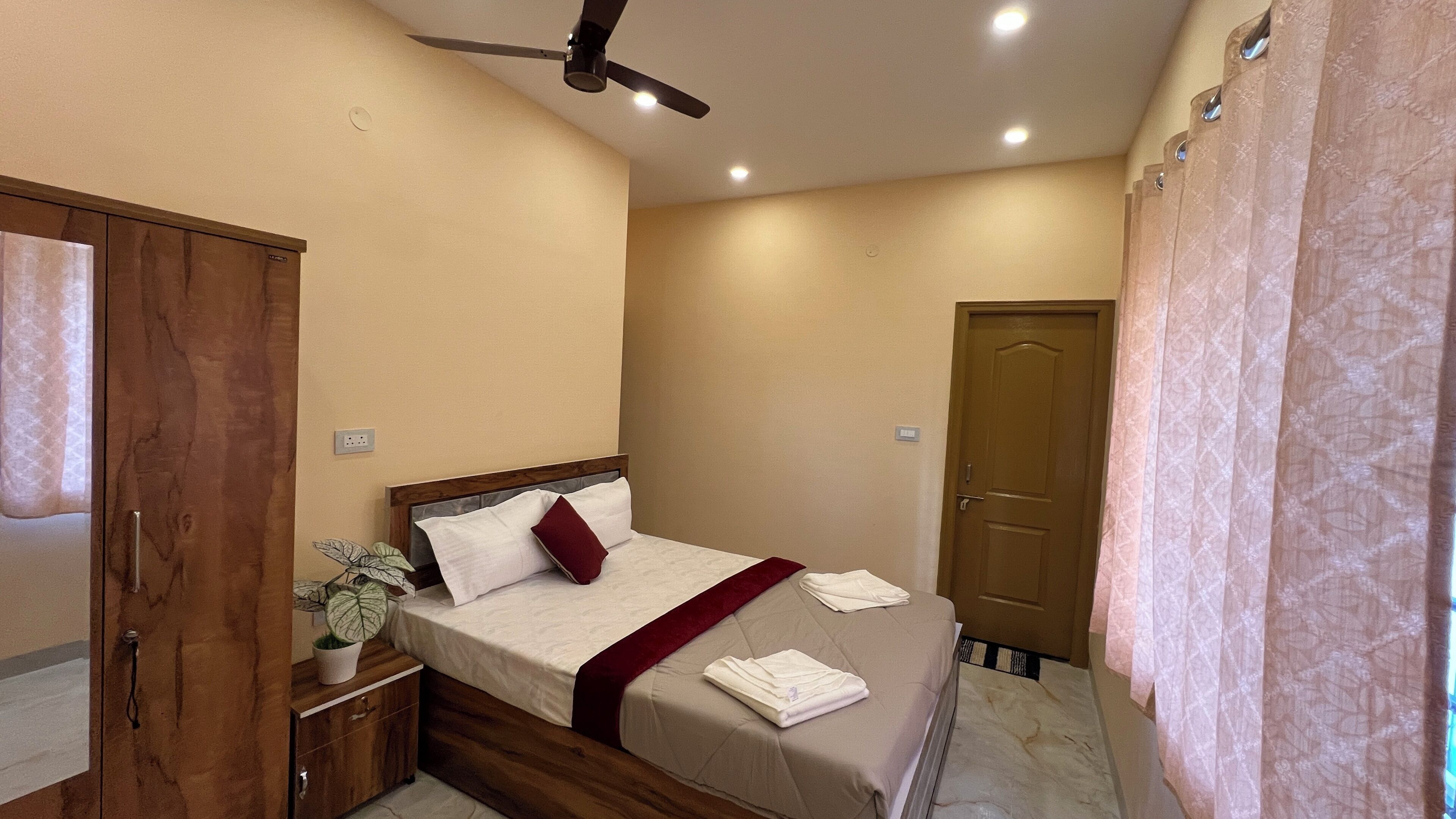 Une chambre à coucher avec un lit, une table de chevet, une plante, un ventilateur de plafond et une porte.