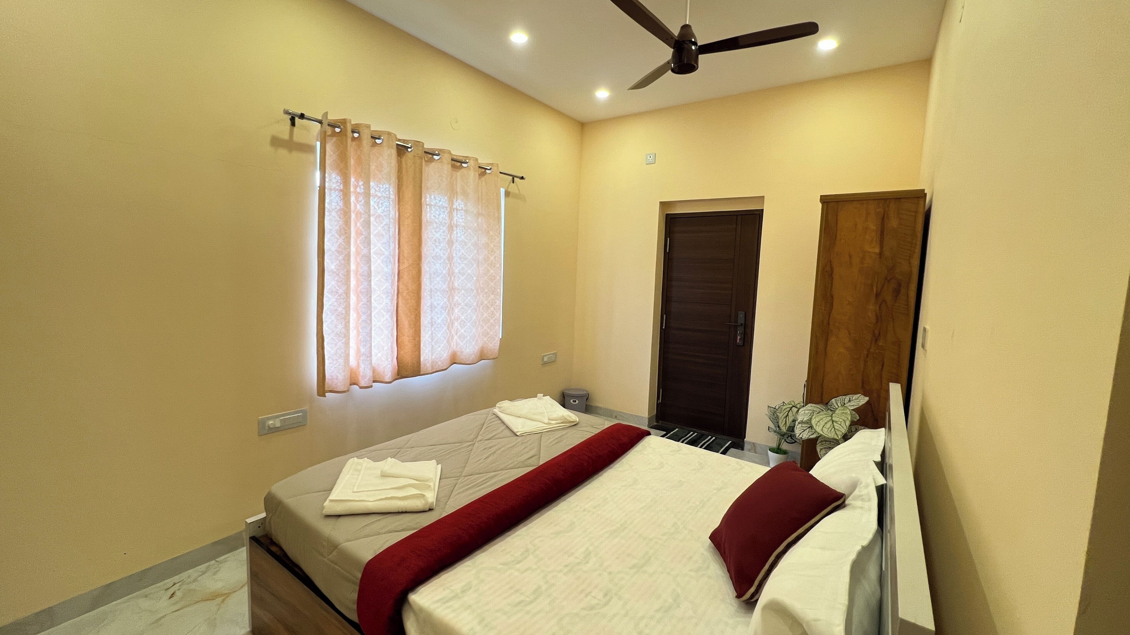 Une chambre à coucher avec un lit, un ventilateur de plafond, une fenêtre avec des rideaux, une porte et une petite poubelle.