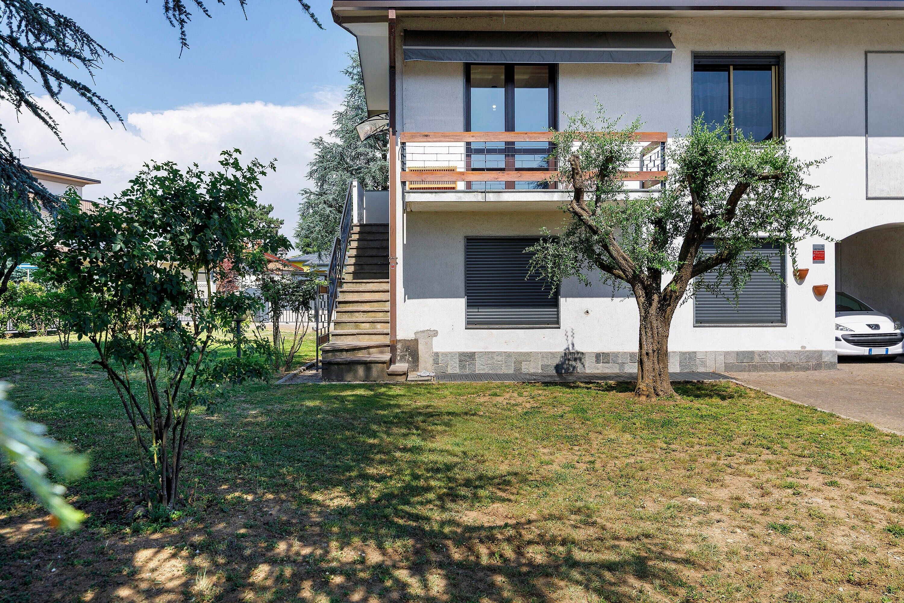 A modern two-story building with a balcony, a tree in the front yard, and a parked car.