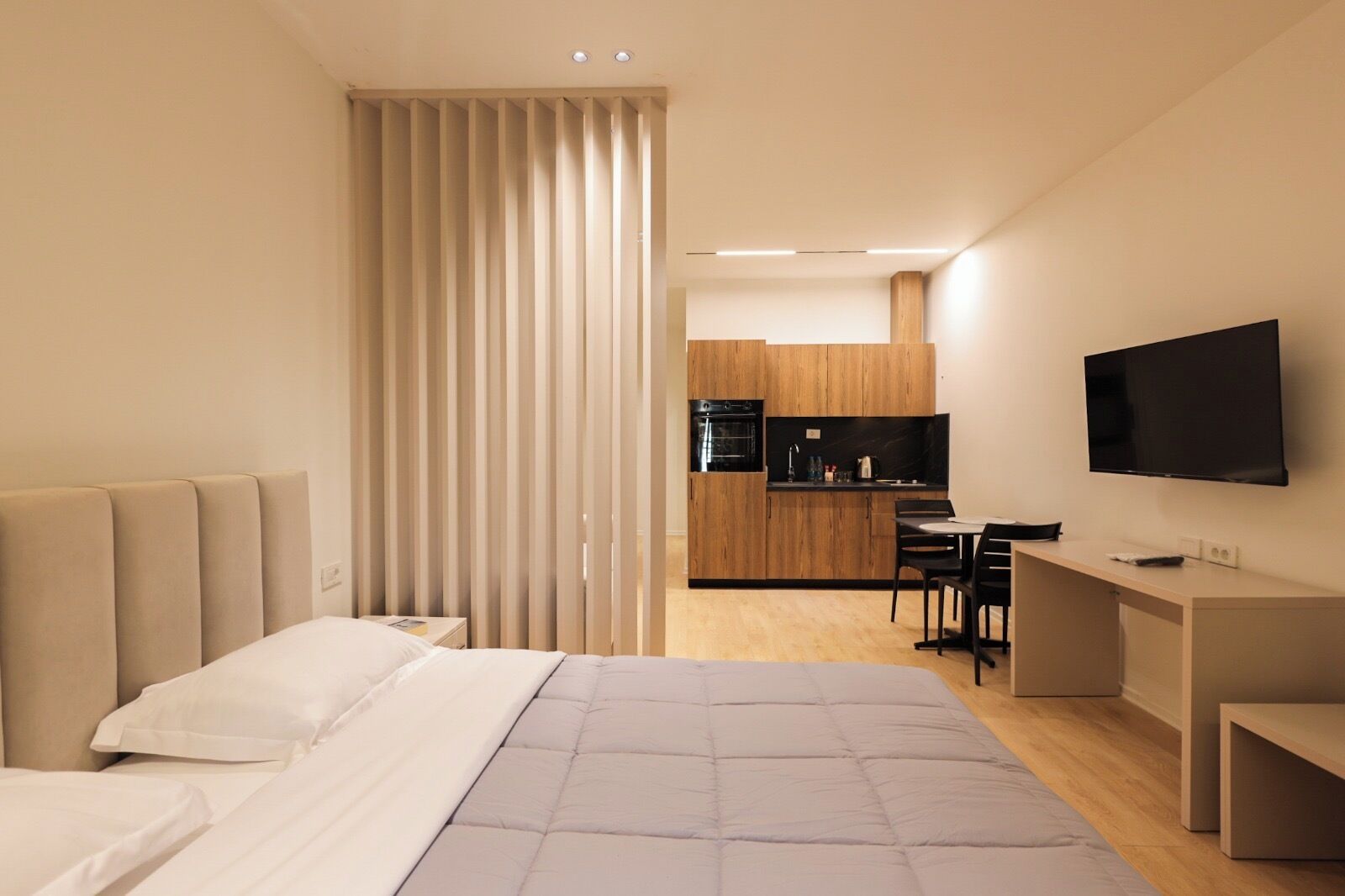 A modern hotel room with a bed, a desk, a TV, and a kitchenette.