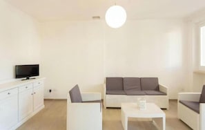 Ein modernes Wohnzimmer mit Sofa, zwei Sesseln, Couchtisch, Fernseher auf einem Schrank und einer Pendelleuchte.