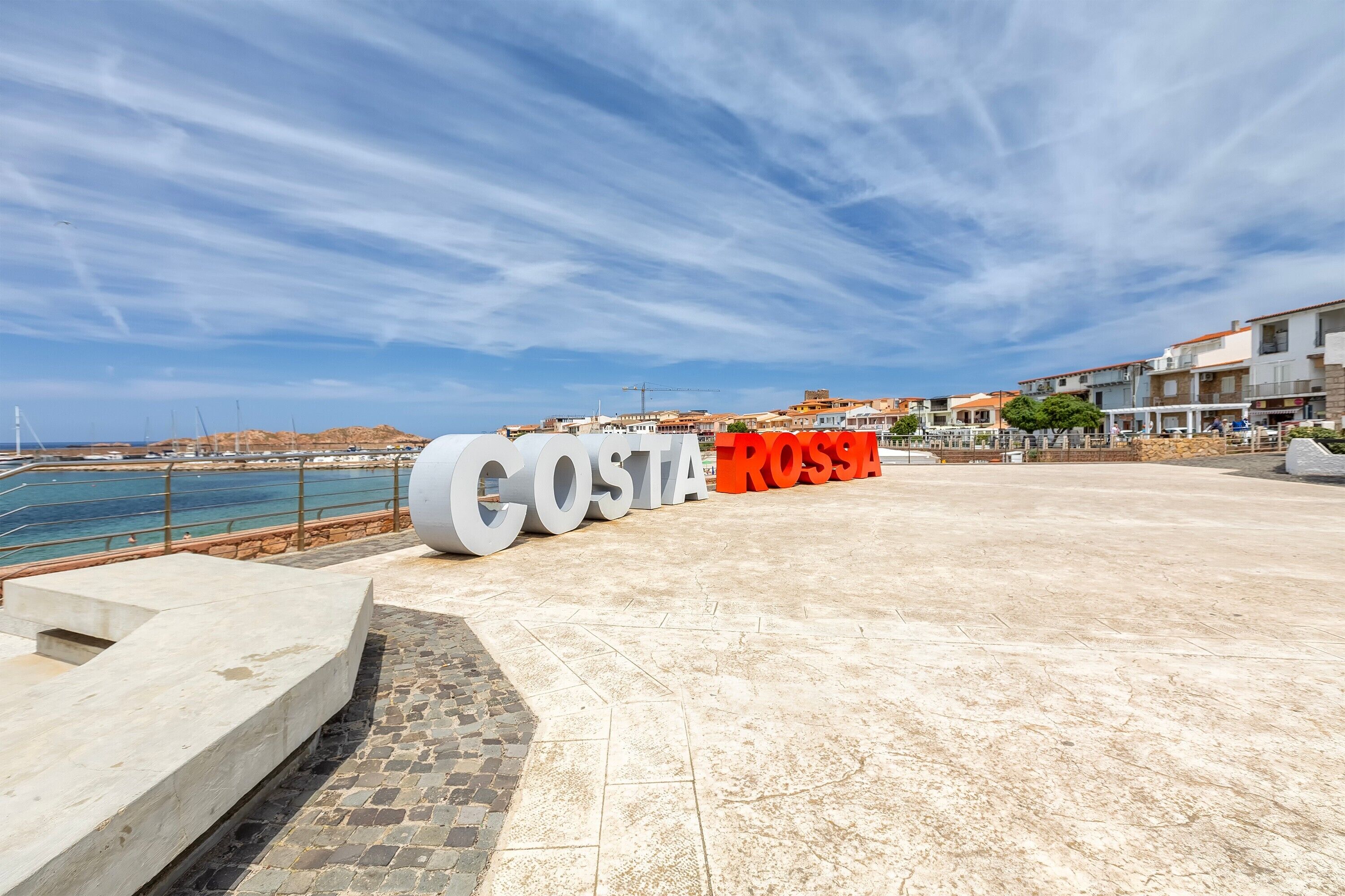A coastal promenade with large letters spelling 'COSTA ROSSA' and a view of the sea and buildings in the background.
