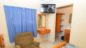 Ein kleines Zimmer mit einem braunen Stuhl, einem Bett mit einer blau-weißen karierten Tagesdecke, einem an der Wand montierten Fernseher, einem Holzregal und einem Spiegel.