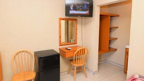 A room with a TV mounted on the wall, a desk with a chair, a refrigerator, and a shelving unit.