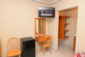 A room with a TV mounted on the wall, a desk with a chair, a refrigerator, and a shelving unit.