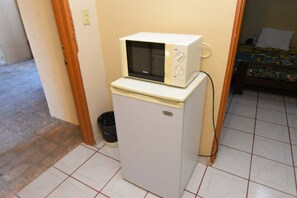 A compact kitchenette with a microwave on top of a refrigerator, a small trash bin, and a bed with patterned bedding in the background. - San Felipe Studio Five (San Felipe)