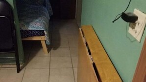 Un couloir étroit avec un banc en bois, un lit avec une literie à motifs et une prise électrique au mur.