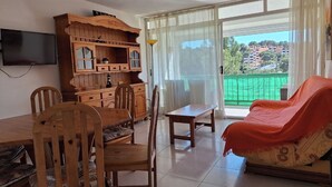 Ein Wohnzimmer mit Esstisch, Stühlen, einer Couch, einem Fernseher und einem großen Fenster mit Blick auf Gebäude und Grünflächen.