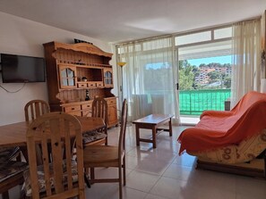 A living room with a dining table, chairs, a sofa, a TV, and a large window with a view of buildings and greenery. - Self-contained apartment with swimming pool 2 steps from the sea (Salou)