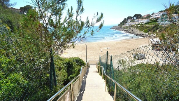 Ein Küstenweg führt zu einem Strand mit freiem Blick auf das Meer und Häusern im Hintergrund.