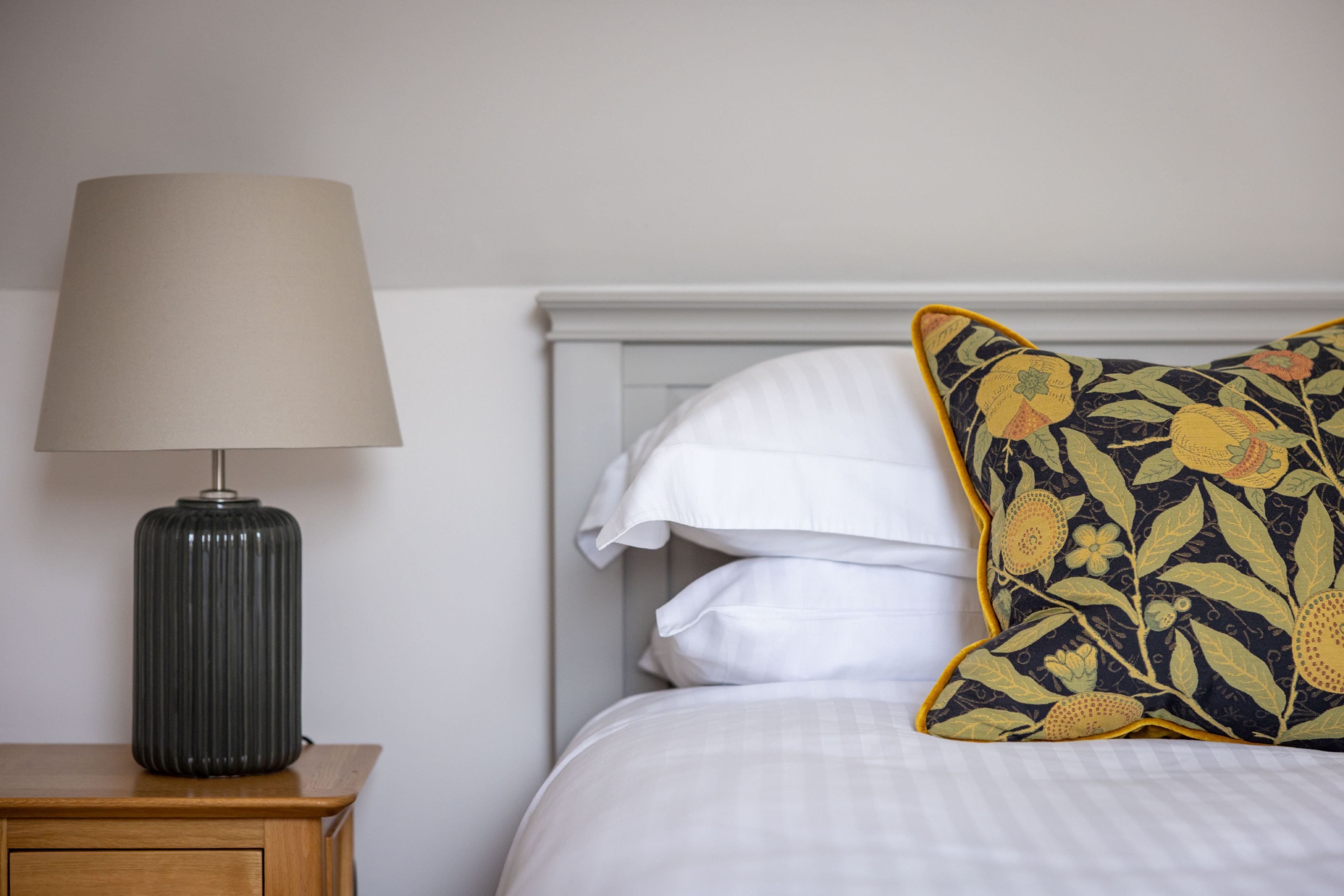 A bed with white bedding, a dark lamp on a wooden nightstand, and a floral pillow.