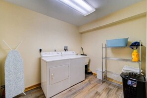 A laundry room with a washing machine, dryer, ironing board, and shelves with cleaning supplies.