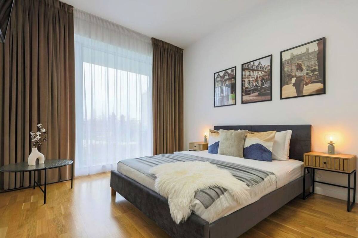 A bedroom with a large bed, two bedside tables, a nightstand, a window with curtains, and three framed pictures on the wall.