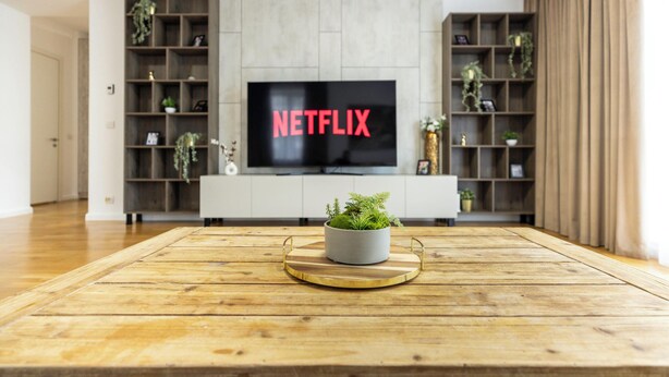 Uma sala de estar com uma mesa de centro de madeira, uma televisão exibindo 'NETFLIX' e um móvel de entretenimento branco com prateleiras.