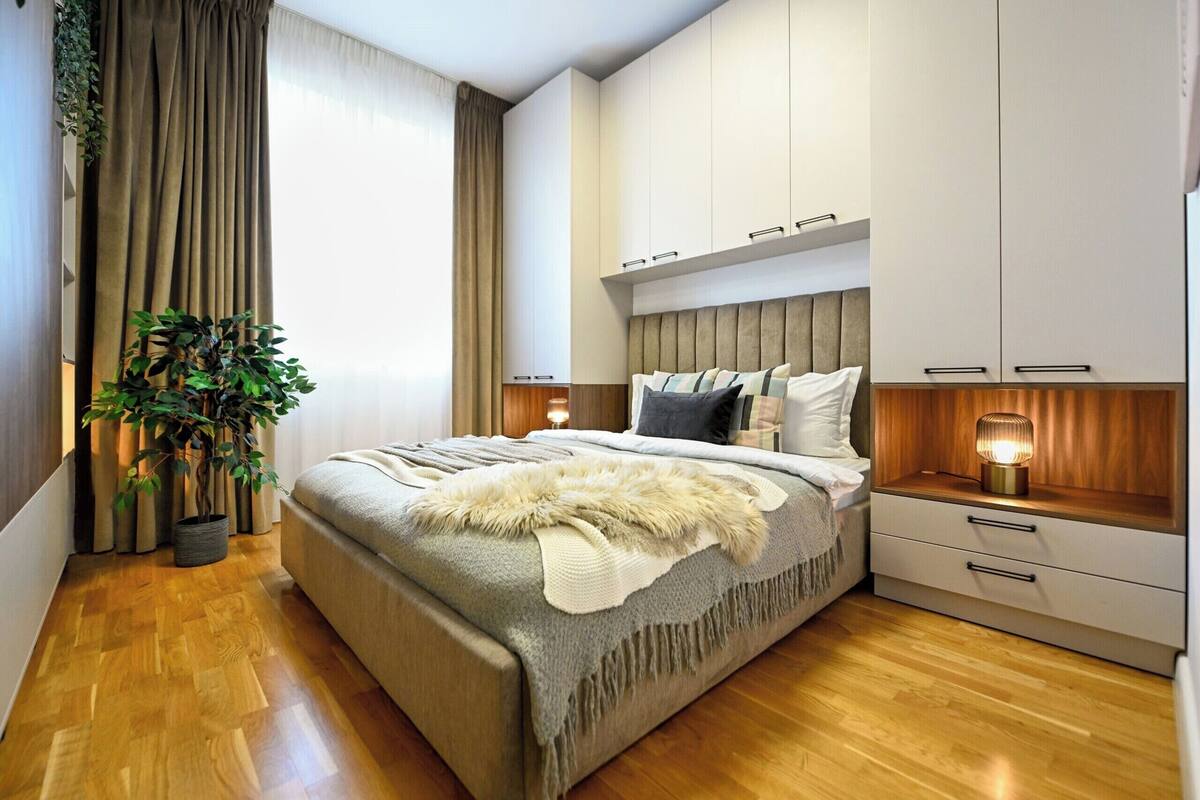 A modern bedroom with a large bed, built-in wardrobe, bedside tables, and a potted plant.