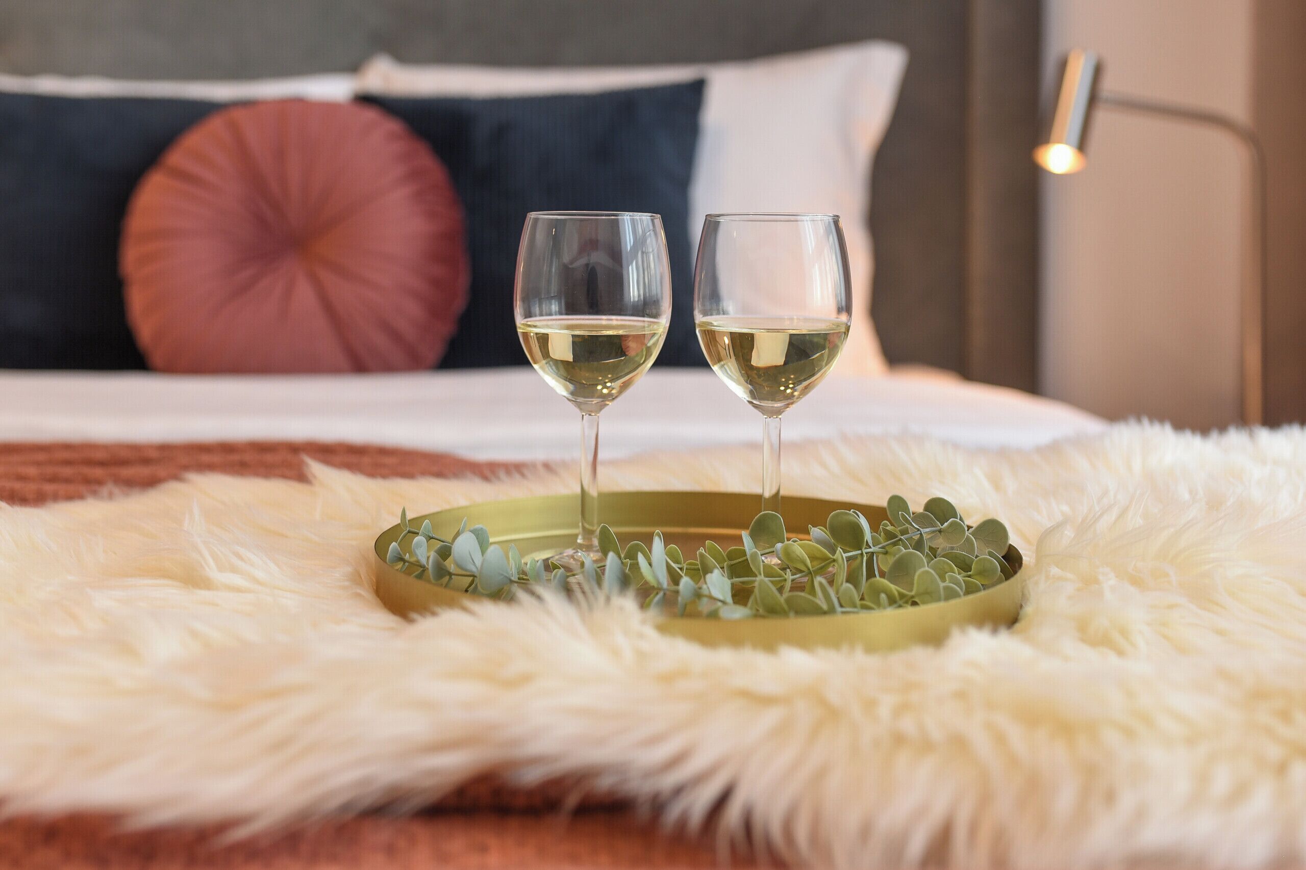 Dos copas de vino blanco sobre una bandeja con hojas verdes, sobre una superficie blanca mullida en una habitación con cama, almohadas y una lámpara.