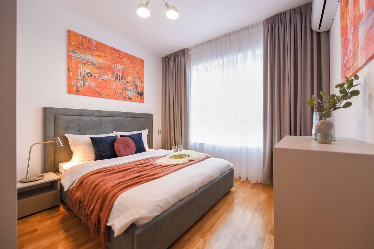 A modern bedroom with a large bed, a nightstand, a bedside lamp, a vase with a plant, and a painting on the wall.