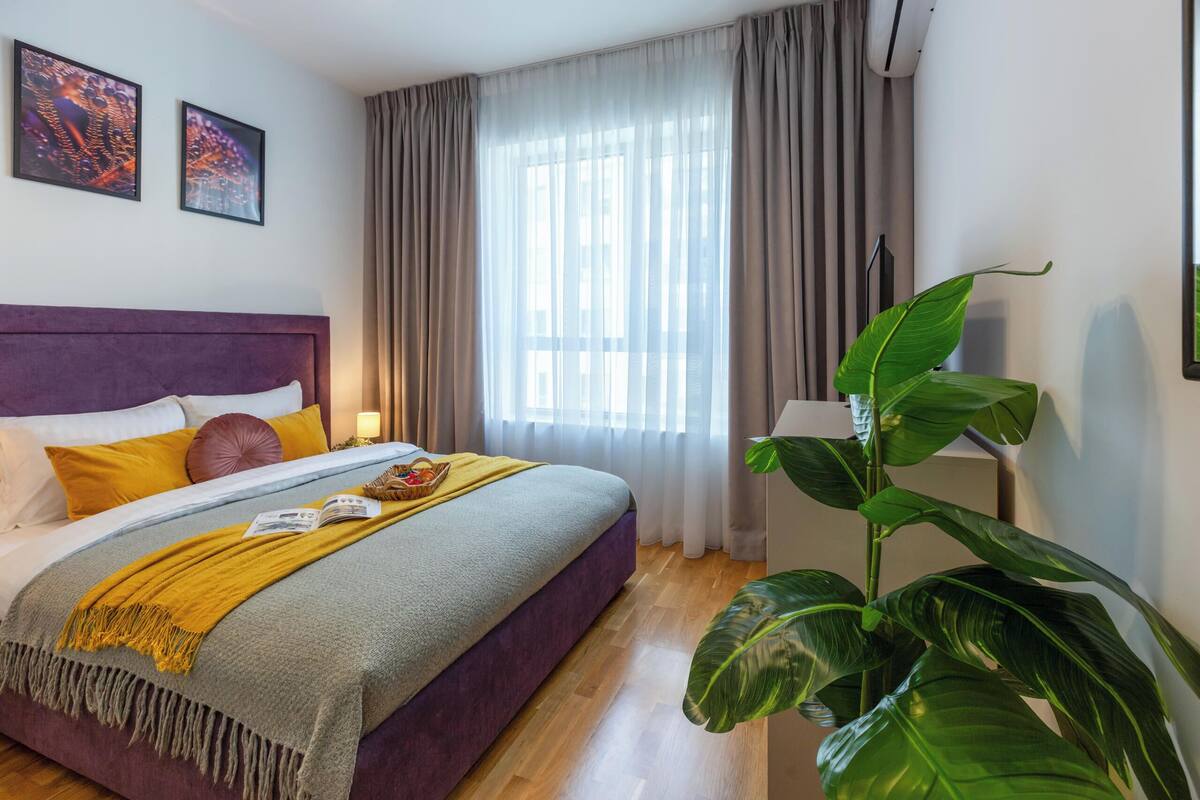A hotel room with a bed, curtains, a plant, and artwork on the wall.