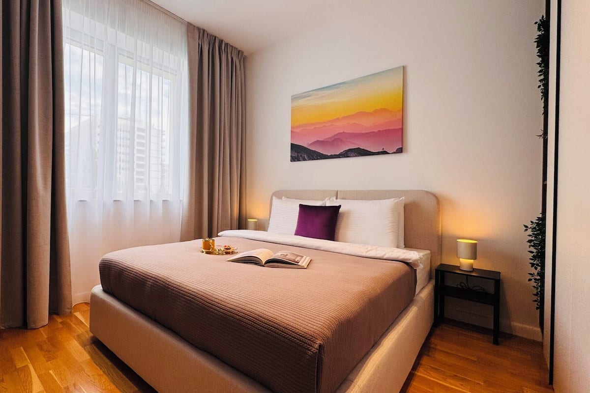A neatly arranged bedroom with a bed, bedside tables, a window with curtains, and a wall-mounted painting.