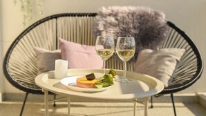 Mesa redonda com prato de comida, dois copos de vinho branco e uma vela em varanda com cadeira de vime e almofadas.