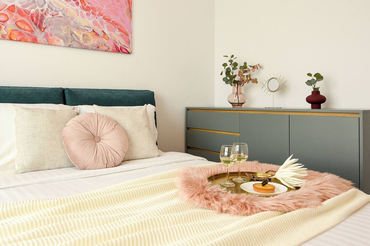 A neatly made bed with a fluffy pink throw, a bedside table with a vase of flowers, and a decorative mirror on a grey cabinet.