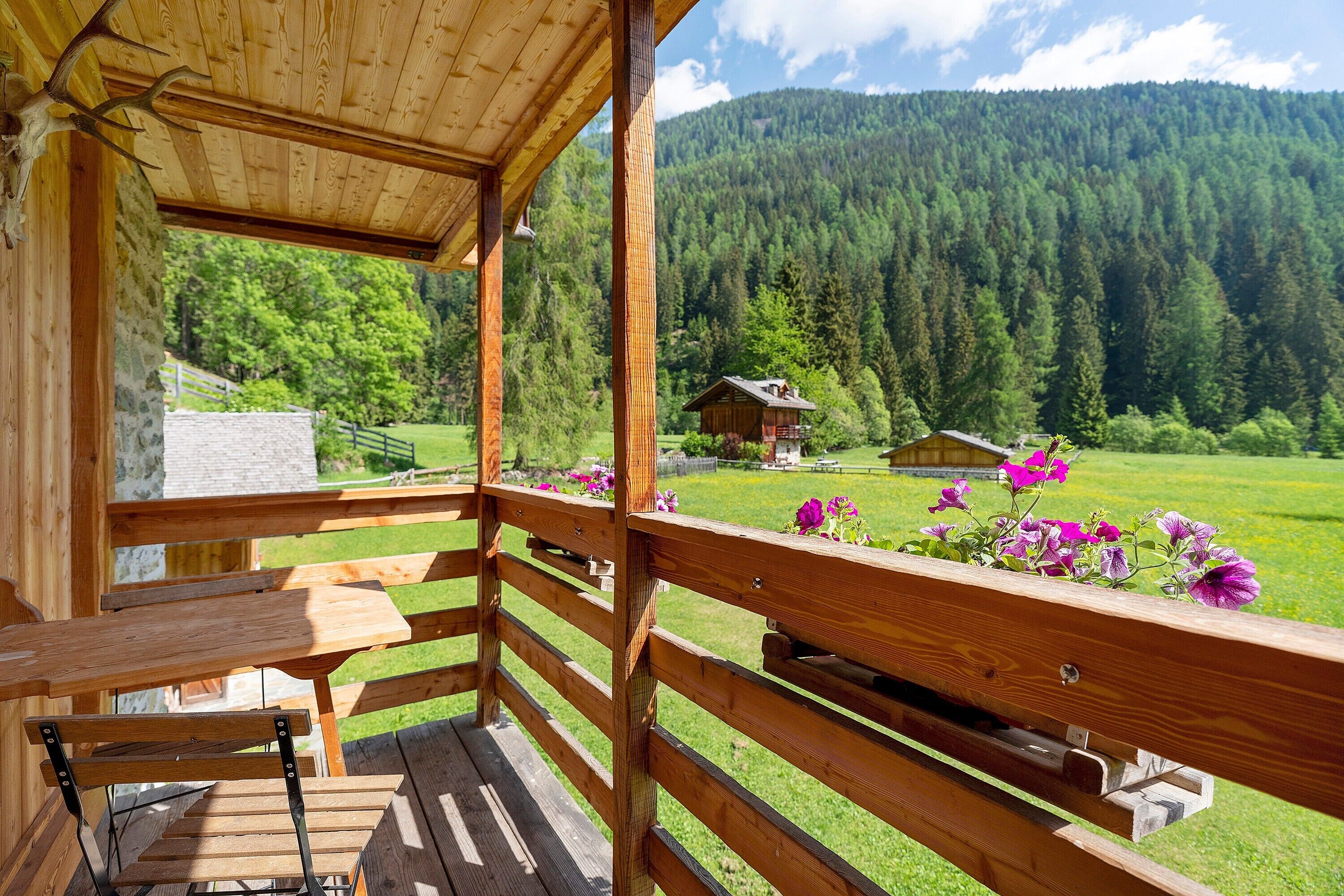 A wooden deck with a bench and a table, overlooking a green field and a forested mountain.