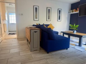 En moderne stue med en blå sofa, et lille træ sidebord, en potteplante og billeder i rammer på væggen.