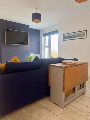 Un salón moderno con un sofá azul oscuro, una mesita de madera, un televisor montado en la pared y una ventana con luz natural.