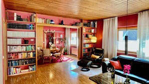 A spacious living room with a wooden floor, a black leather sofa, a dining table with chairs, and a bookshelf filled with books.