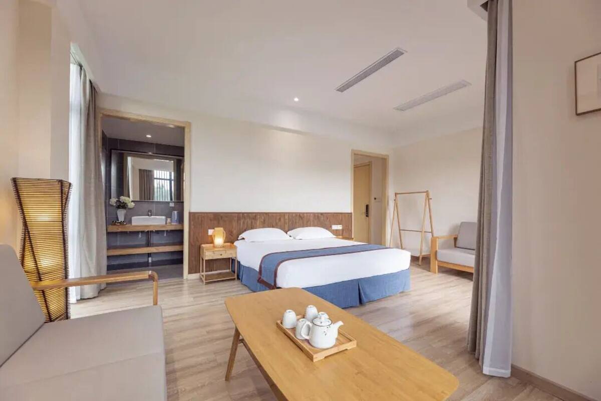 Una habitación de hotel con cama, mesita de noche, lámpara, silla y una mesa de madera con teteras.