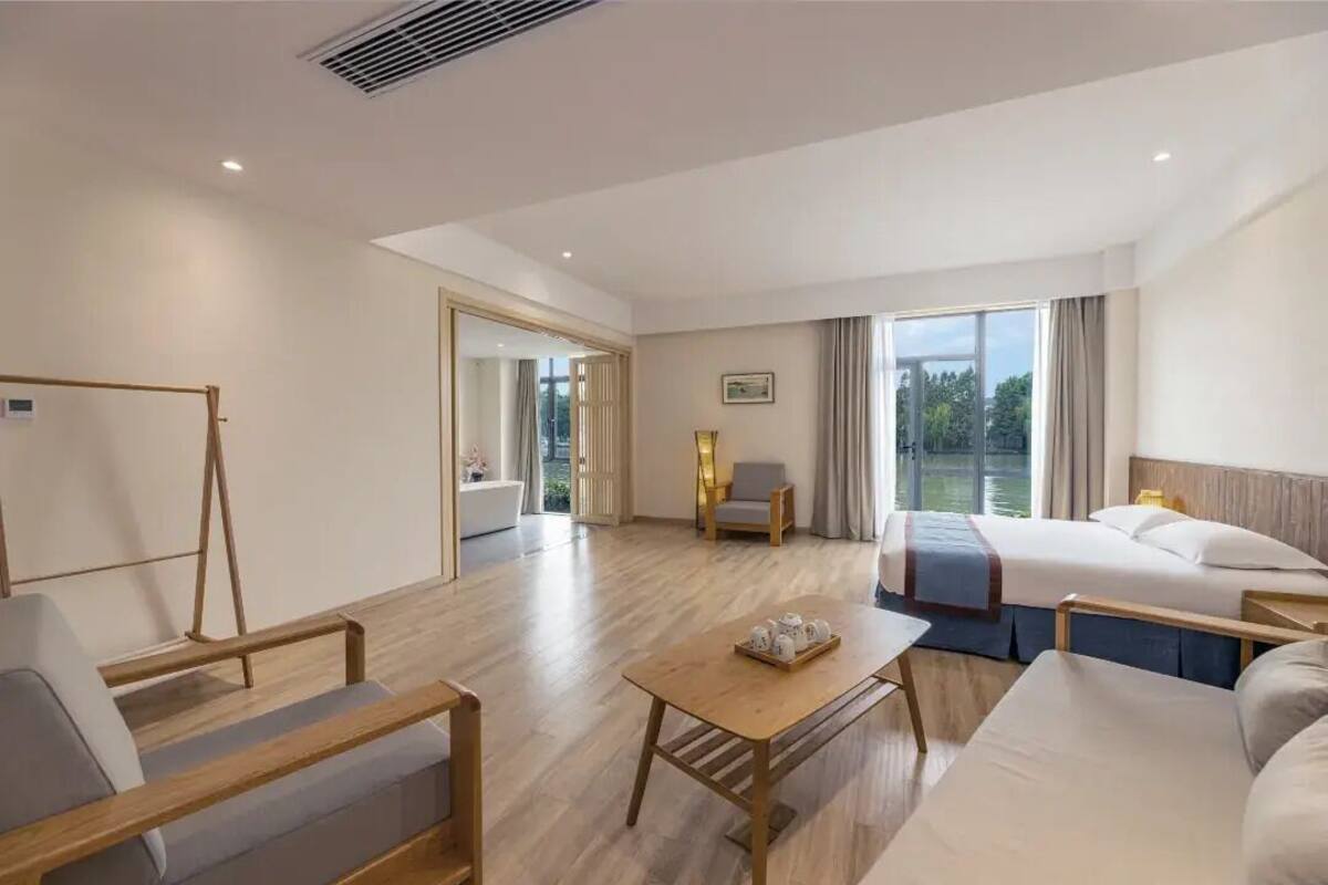 Habitación de hotel con cama, sofá, mesa y silla. Hay una ventana con vistas a árboles y una puerta que da a otra habitación.