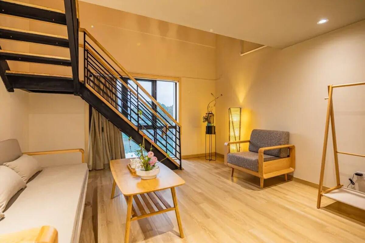 Una sala de estar moderna con una escalera, un sofá, una mesa de madera con un jarrón, una silla y una lámpara de pie.