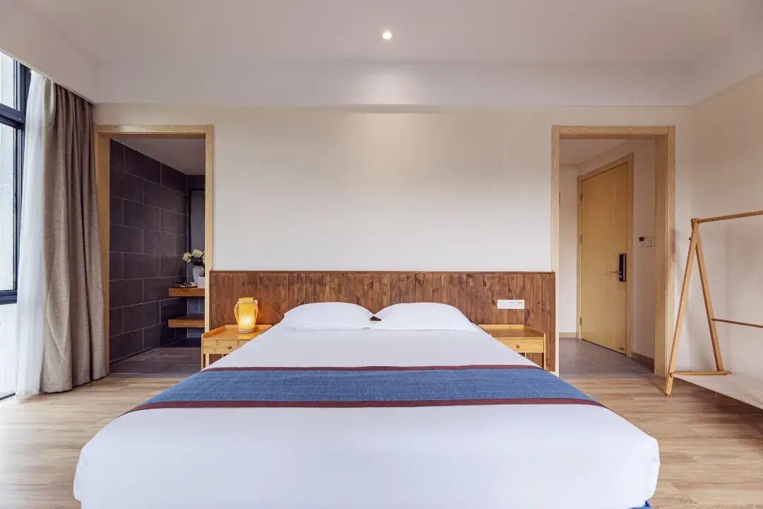 Habitación de hotel con una cama grande, cabecera de madera, mesitas de noche, una lámpara y vistas a través de una puerta abierta.