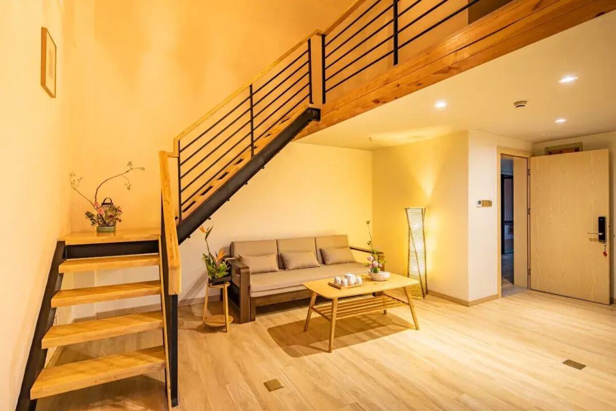 Una escalera con escalones de madera y barandillas negras, un sofá, una mesa de centro y una lámpara de pie en una habitación con suelo de madera y una puerta al fondo.