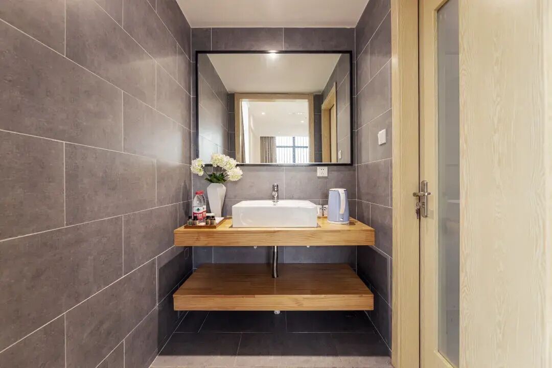 Un baño moderno con paredes de azulejos oscuros, un mueble de tocador de madera, un lavamanos blanco, un espejo y un pequeño estante con artículos de aseo.
