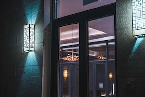 Une entrĂ©e dâimmeuble avec une porte en verre, un luminaire fixĂ© au mur et un intĂ©rieur visible, agrĂ©mentĂ© dâun lustre.