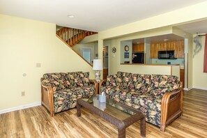 A living room with two floral-patterned sofas, a glass coffee table, wooden flooring, a staircase, and a kitchen area with wooden cabinets. - Marble Winds with a large, oceanfront sundeck with impressive views and built-in (Surf City)