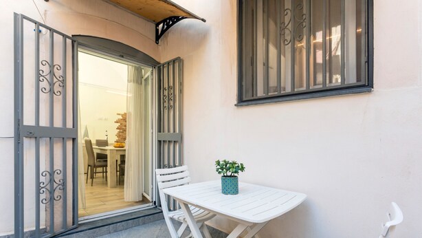 Een terras buiten met een witte tafel en stoelen, een potplant en een uitzicht op het interieur door een open deur.